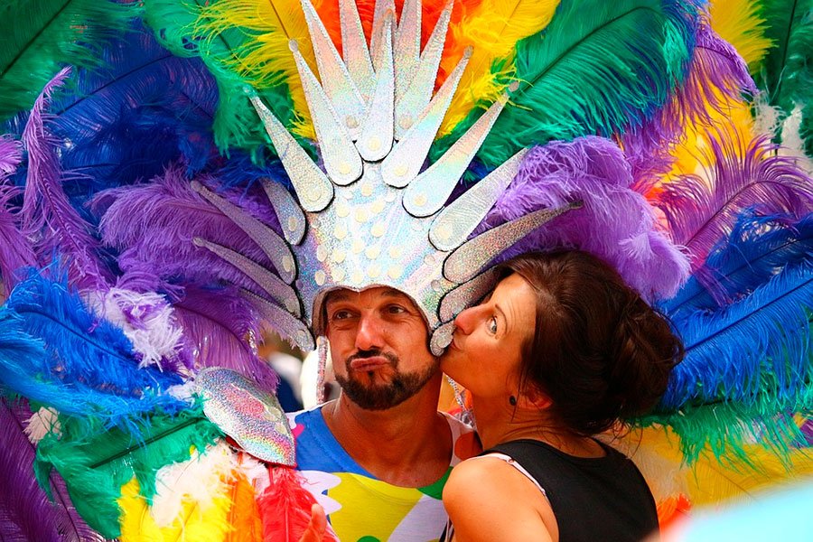 un chico vestido con muchas plumas de colores, y una mujer le besa la mejilla.