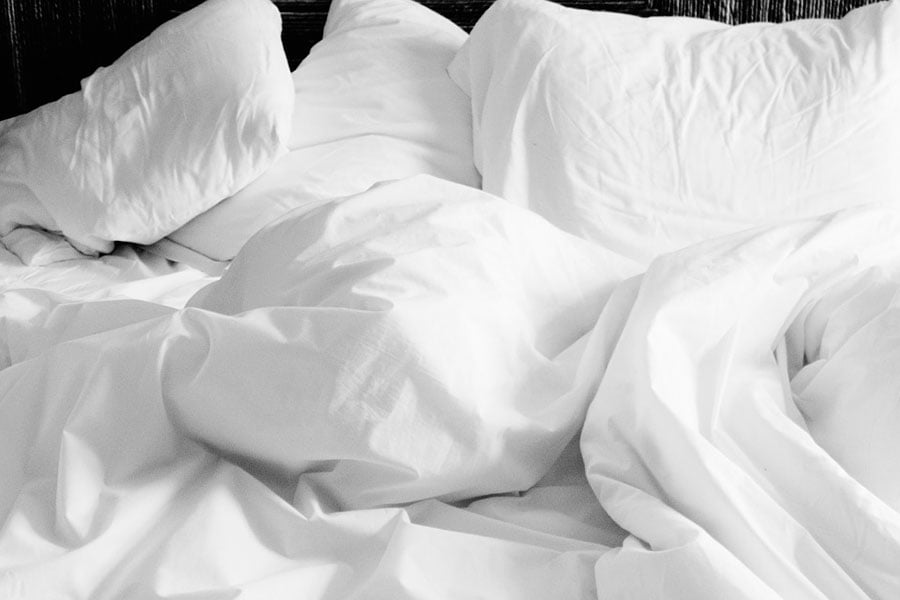una cama sin hacer con un edredon de plumas y una almohada de plumas de oca de diferentes tamaños. Todo es blanco.
