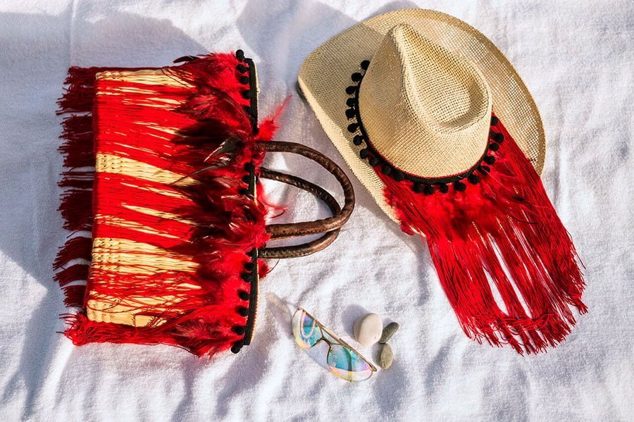 sombrero y bolso de paja decorados con flecos de plumas de color rojo. Están sobre una toalla de color blanco, diría que alguien está en la playa tomando el sol.