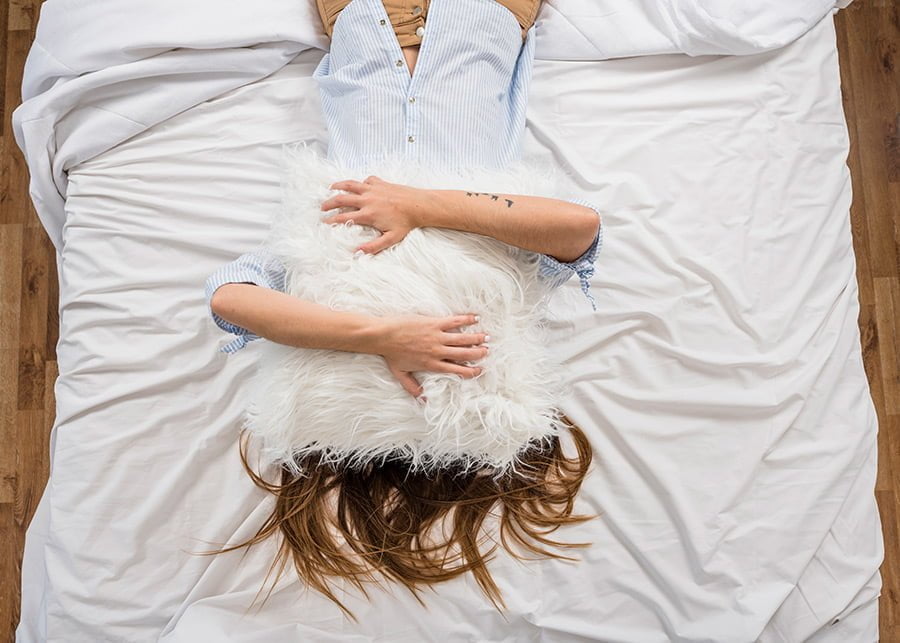 Esta chica está tumbada boca arriba en la cama con un cojín con plumas alrededor de color blanco en la cara. Lo sostiene con sus manos porque está contenta Los cojines adornados con plumas son muy suaves..